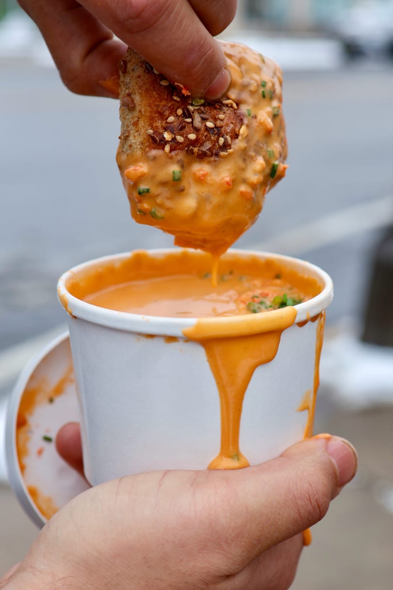A hand dips a piece of bread into a cup of creamy orange soup, dripping down the side.