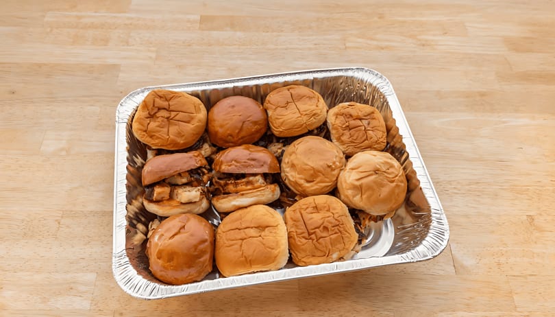 A foil tray filled with a dozen mini sliders, some open to show meat and onions.