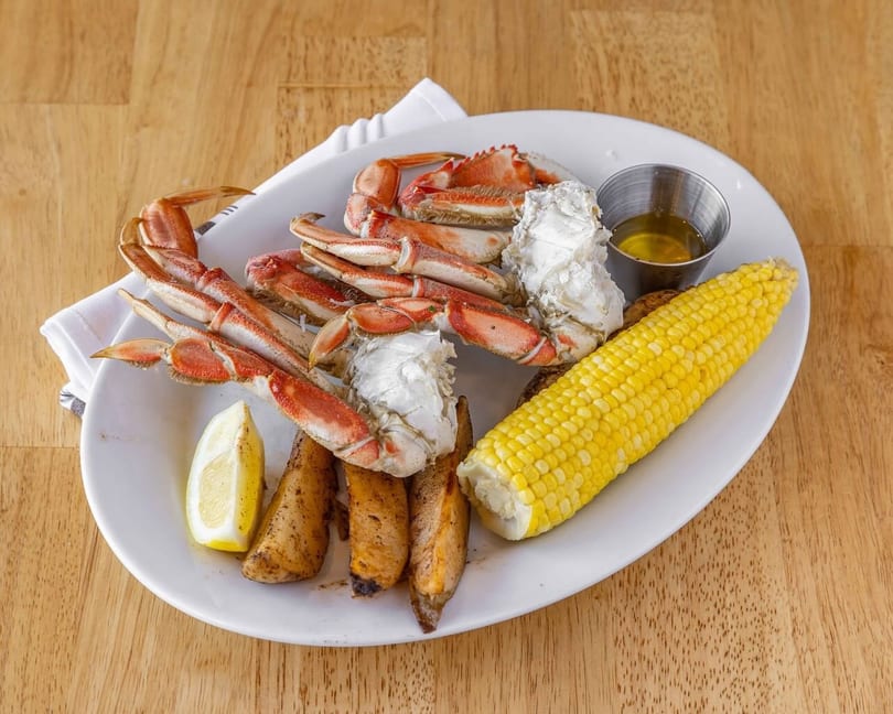 Snow crab legs, corn on the cob, potato wedges, lemon, and butter on a white plate.