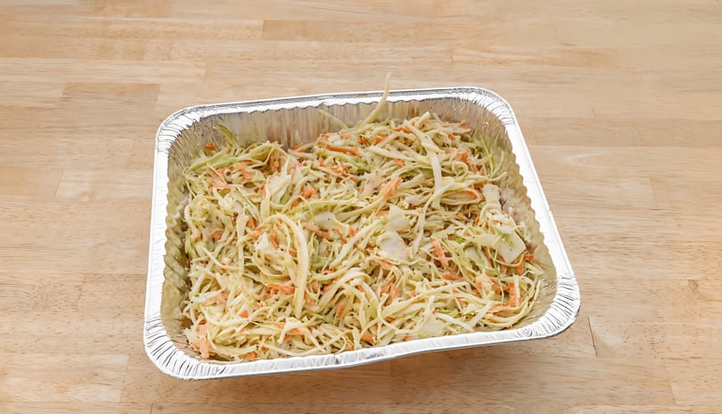 Creamy coleslaw with shredded cabbage and carrots in a large aluminum catering pan on a wooden table.