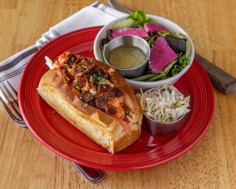 Lobster roll with bacon, coleslaw, and a side salad with watermelon radish on a red plate.