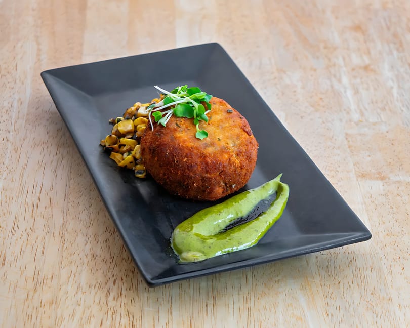 Crispy crab cake with roasted corn, microgreens, and green sauce on a black plate.